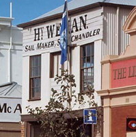 Image: Two elderly women stand in front of a brick-and-corrugated iron two-storey building. The words ‘Hy. Weman. Sail Maker, Ship Chandler’ are painted on the front of the building 