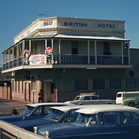 Image: A cream coloured two-storey building with second-floor verandah. Two cars are parked in front of the building and a man stands near its front door