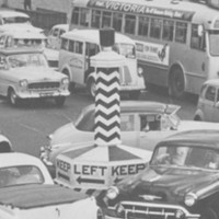 Image: A throng of auto-mobiles of 1940s, 1950s and 1960s vintage circle around a large plinth with the words ‘Keep Left’ painted on it  