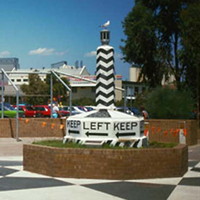 Image: A large plinth painted with an alternating black and white pattern, and emblazoned with the words ‘Keep Left’ and directional arrows at its base, is displayed in a park area made of brick 