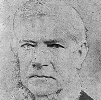 Image: A photographic portrait of a middle-aged man with white hair and large, mutton-chop sideburns