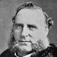 Image: A photographic head-and-shoulders portrait of a Caucasian man with large mutton-chop sideburns wearing late Victorian-era clothing, including an overcoat with fur collar