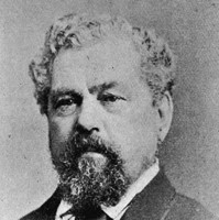 Image: A photographic head-and-shoulders portrait of a middle-aged Caucasian man with curly salt-and-pepper hair and goatee. He is wearing a late nineteenth-century suit and tie
