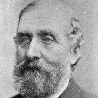Image: A photographic head-and-shoulders portrait of a bearded middle-aged man in a tweed suit with tie