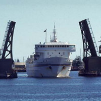 Image: A large, modern steel-hulled ferry passes through an open drawbridge. Part of a wharf is visible in the foreground