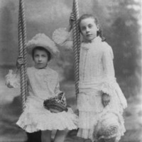 Image: Two young girls pose for a portrait. One sits on a swing, the other stands beside her. They are wearing white, frilly dresses.