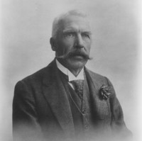 Image: Portrait photograph of a man wearing a suit and tie. He has a large mustache and his hair is white