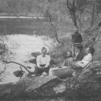 Image: Three women and two boys have a picnic on the banks of a river.