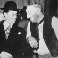 Image:Smiling man in suit and hat next to man in make-up and dirty vest