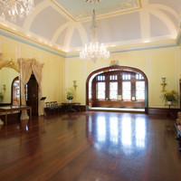 Image: A large, open room with a wooden floor, yellow painted walls, an ornate ceiling, and portraits hanging on the walls