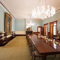 Image: A large room containing a long table with chairs. Two large portraits and a mirror hang on the walls