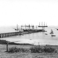 Image: A jetty jutting out into the ocean surrounded by various size sailing ships.