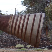 Image: The rusted exhaust section of a rocket mounted on a pedestal near a cluster of gum trees