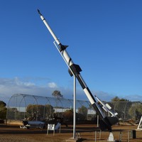 Image: Several different rockets are exhibited on pedestals on a flat, scrubby expanse of ground