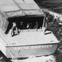 Image: A boat with a group of children sitting in the stern travels along a waterway. Two policemen are visible standing at midships on the starboard side