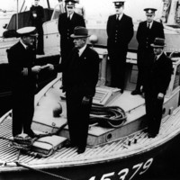 Image: Six men stand on the bow of a boat. One man in a naval uniform passes a paper document to another man in a civilian suit