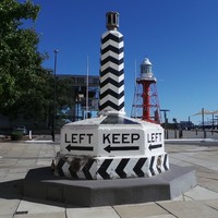 Image: A large plinth painted with an alternating black and white pattern, and emblazoned with the words ‘Keep Left’ and directional arrows at its base. It is displayed on a street corner. A red lighthouse tower is visible in the background