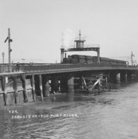 Image: A steam train crosses a bridge that extends across a narrow river. Several buildings are visible along the waterfront in the background