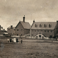 Image: six children, four girls and two boys, walk across a grass paddock in front of a complex of two storey stone buildings with steeply pitched roofs