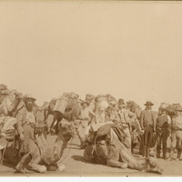 Image: ten camels, all with luggage and saddlebags tied to them with rope, and eight men preparing for an expedition