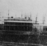 Image: A large paddock, behind which is a large complex of long, single-storey late Victorian-era stone buildings with steam trains parked in front of them. A two-storey building and several ship masts are visible in the distant background