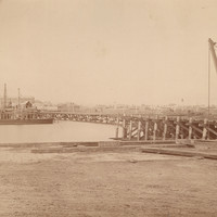 Image: A large iron and wood bridge under construction. Both approaches are nearing completion, while the central span is yet to be started. A barge with cranes is moored in the river near the bridge