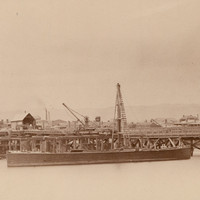 Image: An iron and wooden bridge under construction extends across a narrow river. A nineteenth century sailing vessel and a couple of buildings are visible in the background. A wooden-hulled barge with a derrick is moored next to the bridge