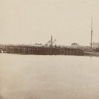 Image: The approaches to a large wooden and iron bridge are in the process of being built. A section of river containing a number of nineteenth century sailing ships are visible in the right foreground