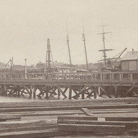 Image: A wooden footbridge extends across a river. A handful of nineteenth-century buildings and sailing ships are visible in the background