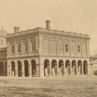 Image: A large, two-storey stone building in Victorian Italianate style. It features open archways on the ground floor, and its central section is topped by a cupola. A group of men stand in front of the left side of the building