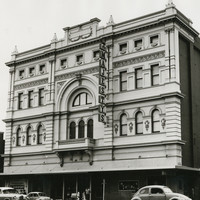 Image: a four storey building with highly decorative facade and plain brick sides. A large arch surrounds a balcony in the centre of the facade and the entrance is protected by a verandah. 1960s era cars are parked outside.