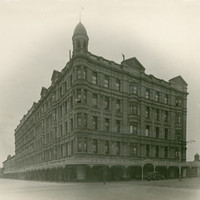 Image: a huge five storey hotel building on the corner of two wide dirt streets. The building has a rounded corner topped with a cupola. 