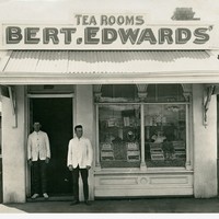 Image: Two men stand in the doorway of a small shop building. A sign on the roof of the shop reads ‘Tea Rooms, Bert.Edwards’