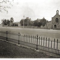 Image: A large stone church and stone house stand adjacent to one another next to a dirt street. Part of an iron fence is visible in the immediate foreground