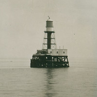 Image: A metal lighthouse with a large wooden support platform stands surrounded by water. Large steam- and sail-powered ships are visible in the distant background