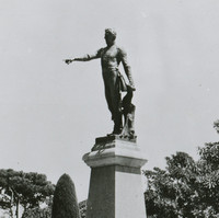 Image: A statue of a man upon a plinth surrounded by formal gardens. His arm is outstretched with his finger pointing.