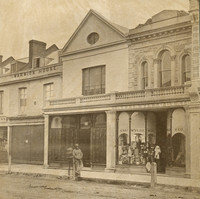 Image: A man and a boy in 1870s clothing stand outside a row of two storey terraced shops. The man is leaning on a post and the boy stands behind him. 