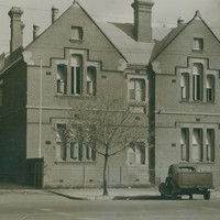 Image: A 1930 era car is parked in front of a plain two storey brick building with gable roofs.