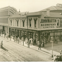 Image: a city department store, mostly two storeys but with one three storey section. Men and women in early 20th century clothes are peering in the plate glass windows at the displays. 