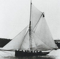 Image: A small, single-masted sailing vessel with a cutter rig coasts off a nearby shoreline. A crew of approximately a dozen men are visible standing along the vessel’s length 
