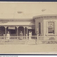 Image: A small group of men and women stand in front of a one-storey stone house with a covered verandah. A white picket fence separates the house from a dirt street