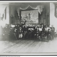 Image: Photograph of members of the German Club Band with their instruments. The picture behind on stage depicting a German rural scene.