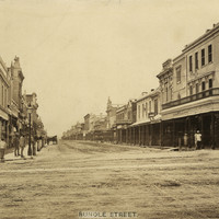 Image: a wide dirt city street, lined with shops, with a single horse drawn vehicle parked on the left of the street. Men are posing for the photograph on both sides of the road outside of shops. 