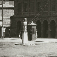 Image: An intersection of two streets in an urban area. A plinth with the words ‘Keep Left’ stands in the centre of the intersection. Two early twentieth century cars are parked in the background 