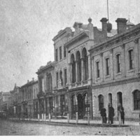 Image: a city street lined with two and three storey buildings. In the foreground is a corner hotel with patrons standing outside.