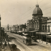 Image: a city street with a number of pedestrians and a horse drawn bus. Towards the centre of the view is an ornate two storey building with a dome. 