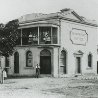 Image: Men, women and children gather in front and on the balcony of a small hotel, painted white with a corner door and the name "Hampshire Hotel" surrounded by a decorative border on the side.