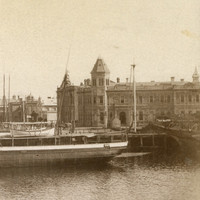 Image: A nineteenth-century steamship with twin funnels and two masts moored at a wharf on the waterfront of a port town. A handful of large nineteenth-century warehouses and other buildings are visible along the waterfront