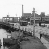 Image: A tram and four pedestrians cross a bridge across a river in a port. A complex of nineteenth-century buildings and a wooden-hulled hulk are visible in the background