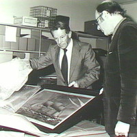 Image: Black and white photograph of two men in suits. They are standing around a just unwrapped painting of a ship